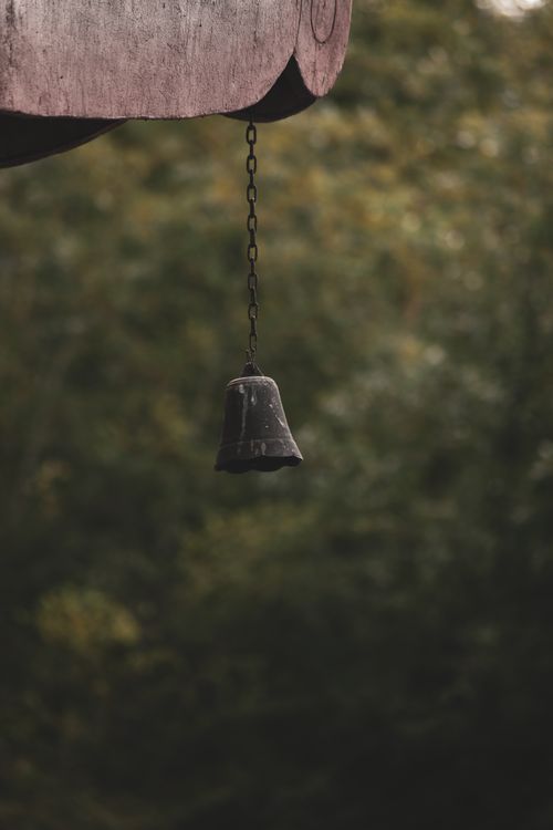 Free A small metal bell hanging outside with a chain, set against a blurred green forest background. Stock Photo
