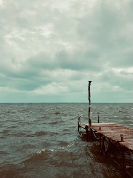 A rustic jetty extends into a stormy sea under a cloudy sky, capturing the mood of a coastal seascape.