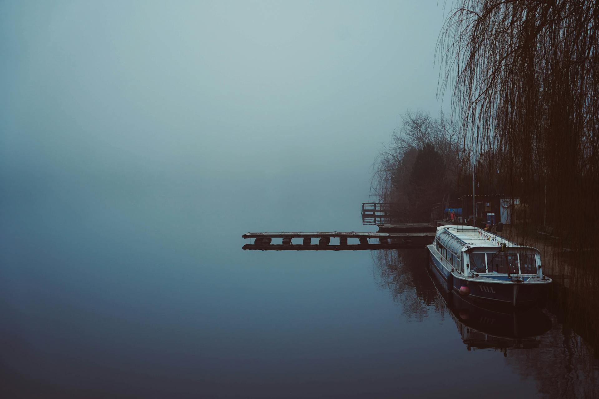 Misty lake scene with a lonely boat docked at a wooden pier on a foggy day.