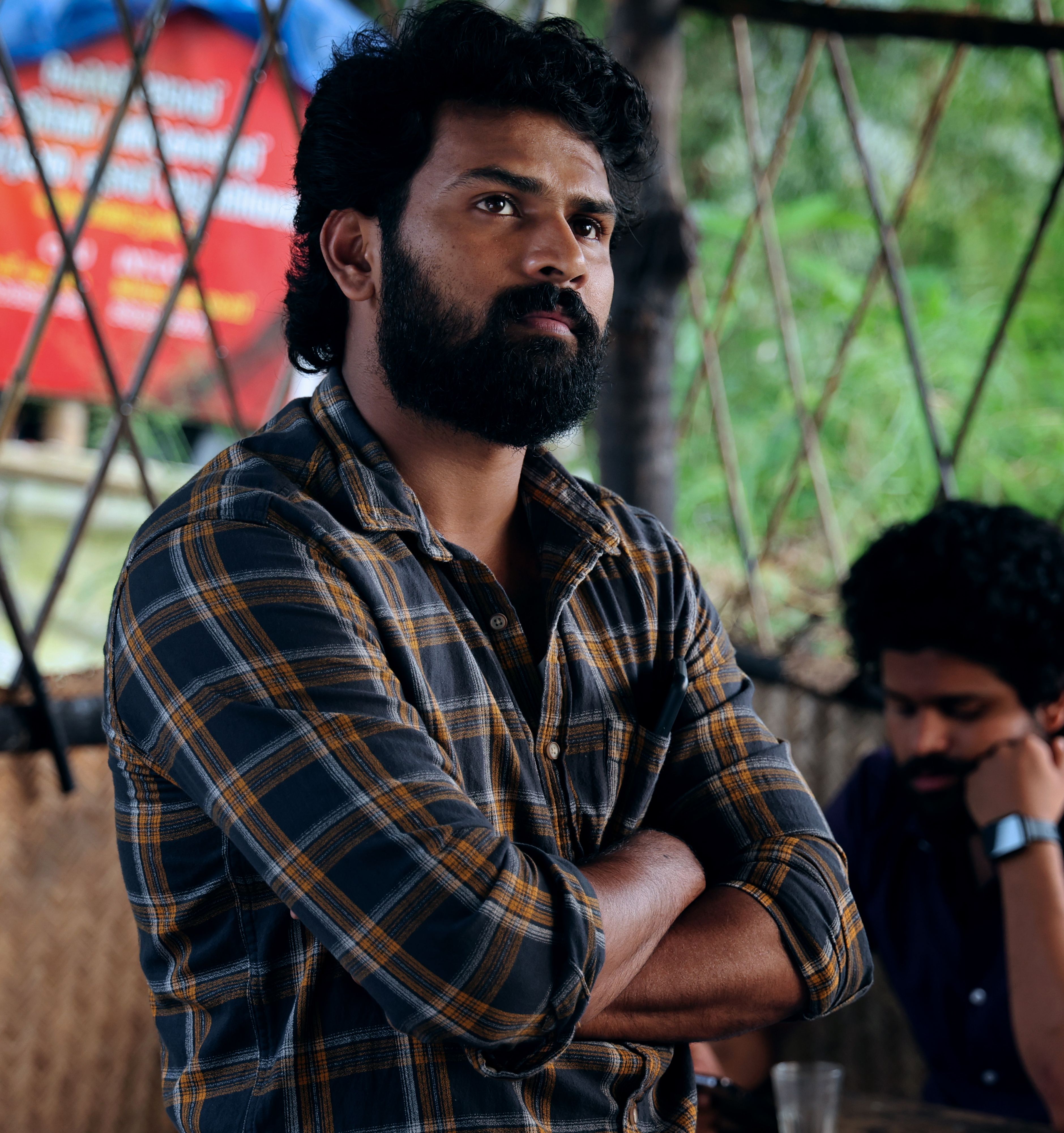 Man with a thick beard and plaid shirt standing indoors with arms crossed.