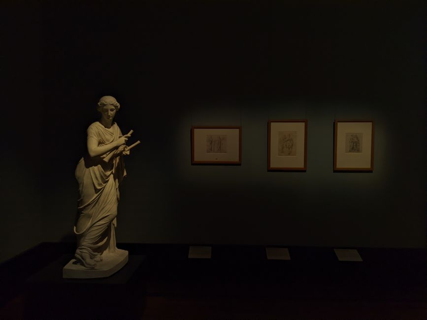 A marble statue and framed drawings in a dimly lit museum exhibition in Berlin.