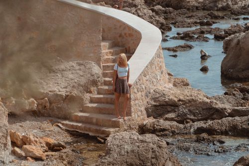 Free Blonde woman standing on stone stairs by the rocky seashore during summer vacation. Stock Photo