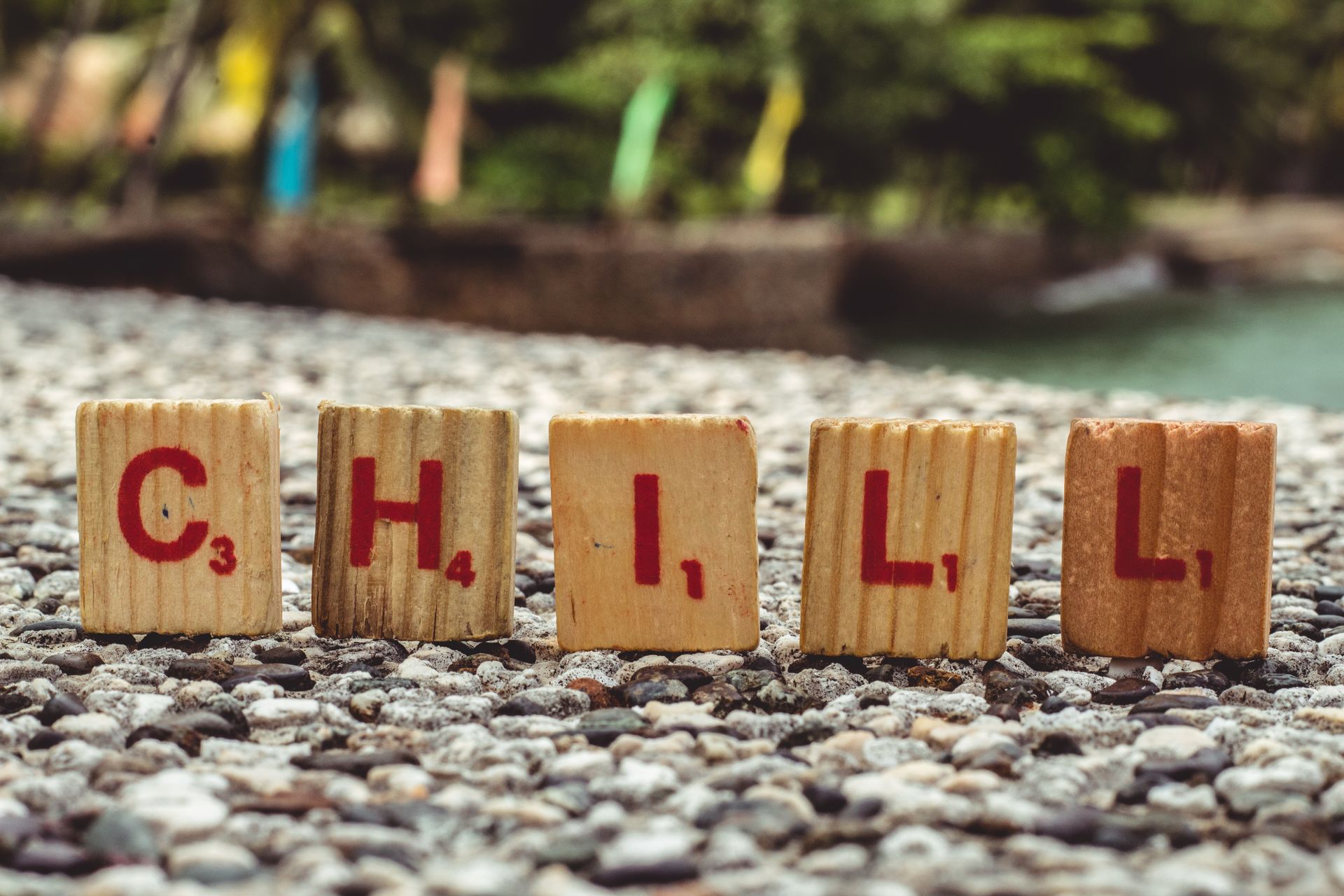 Wooden Scrabble tiles form the word 'CHILL' on a pebbled surface in a garden setting.