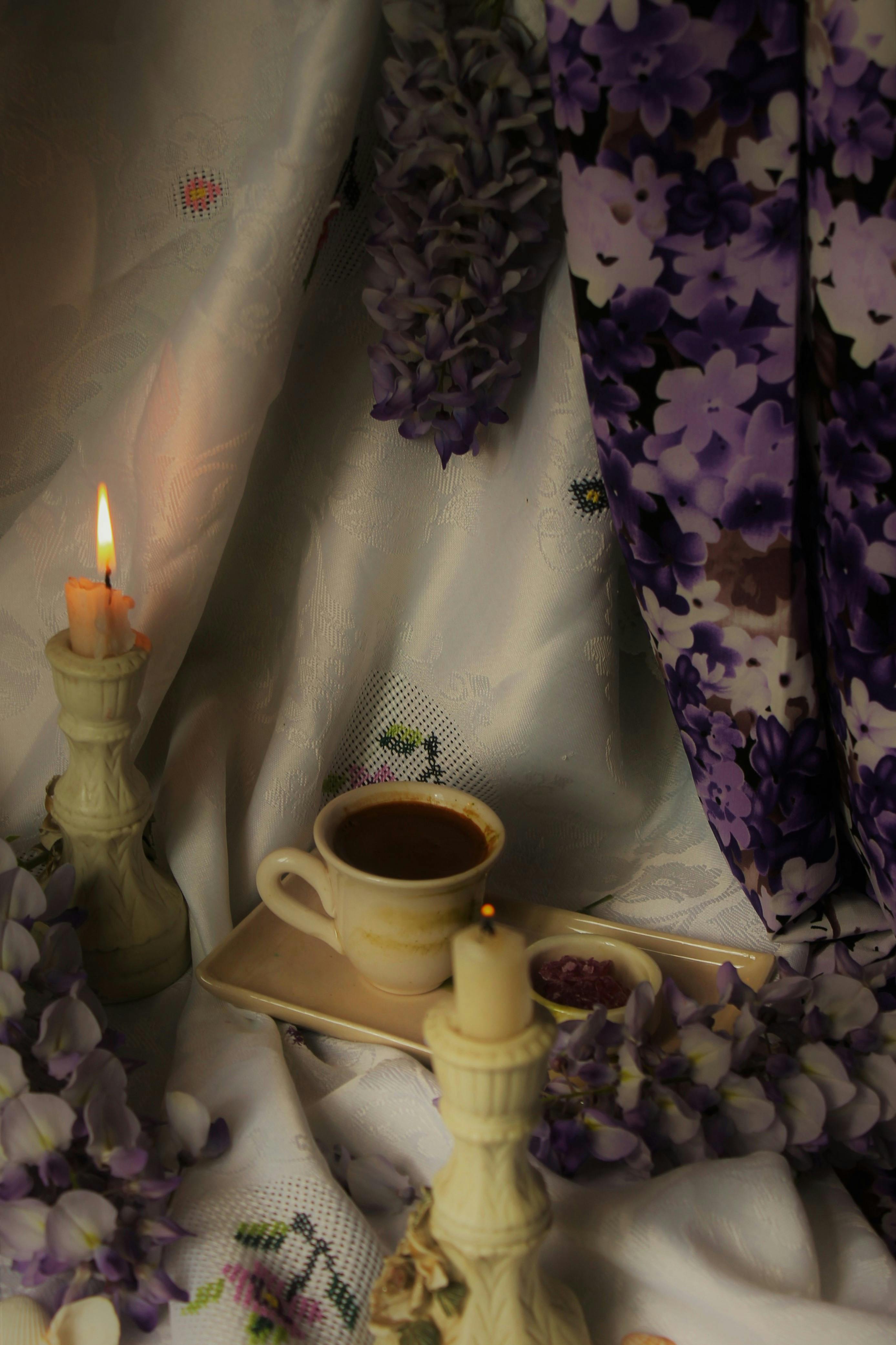 Elegant composition of coffee cup with candles and lilac flowers on white fabric, creating a romantic ambiance.