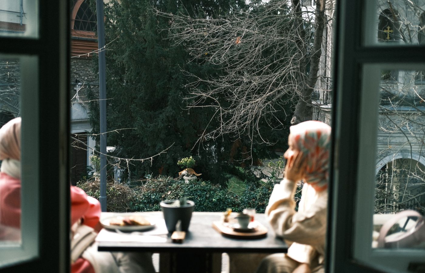 Two women enjoy coffee on a cozy balcony, surrounded by urban greenery.