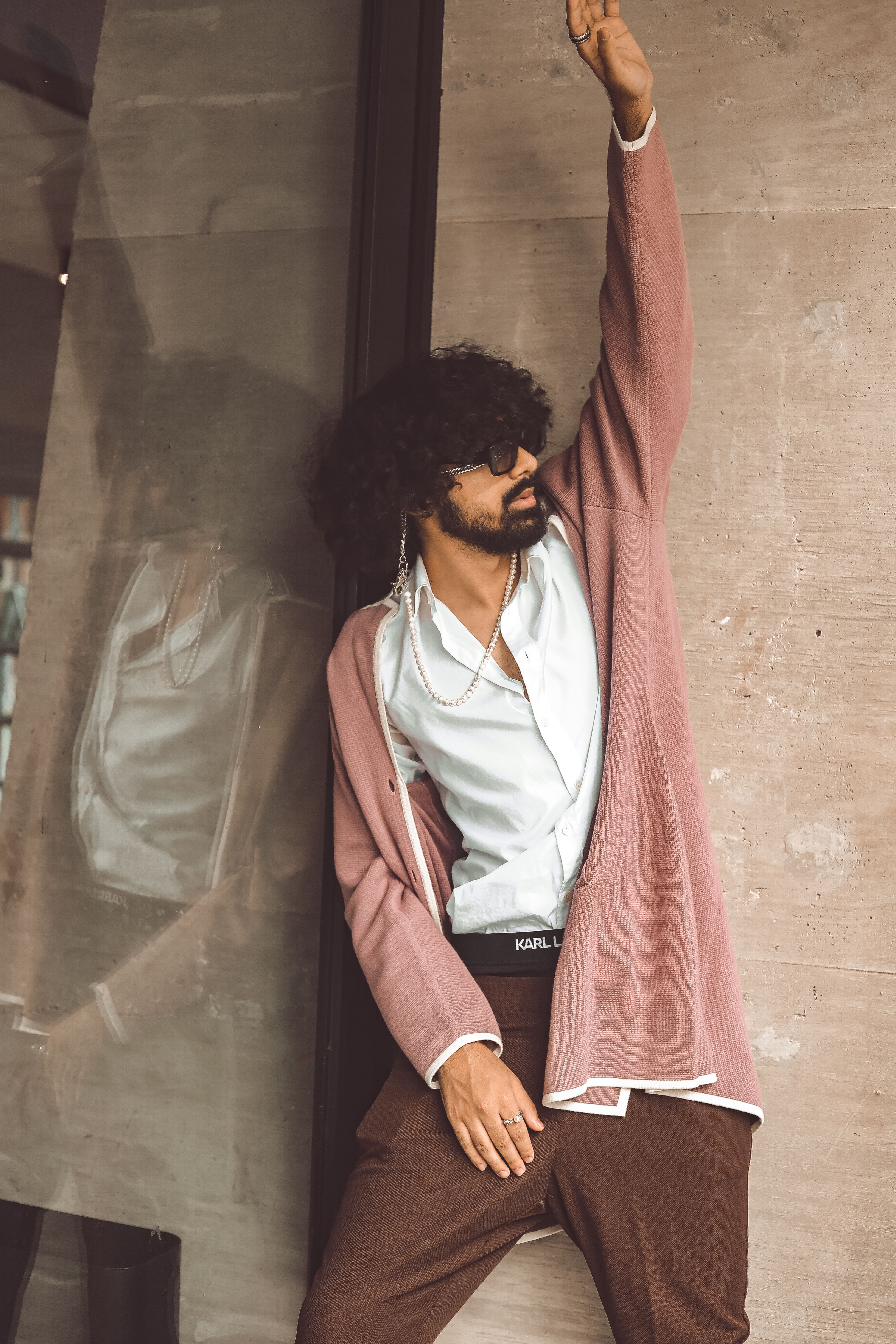 Fashionable man in a pink jacket poses against a concrete wall with an arm raised, showcasing contemporary style.