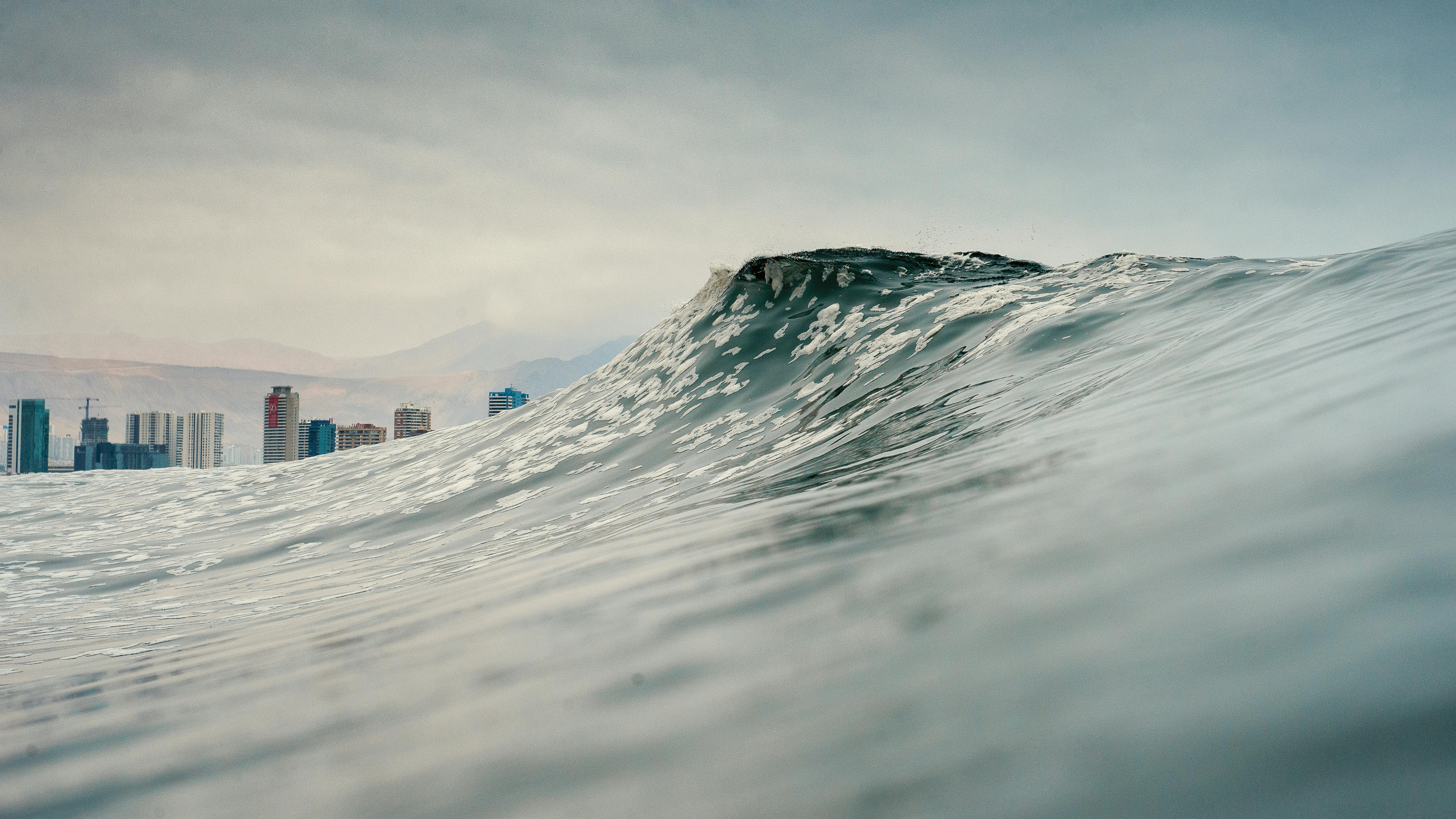 Dramatic ocean wave with Iquique city skyline in the background under a cloudy sky.