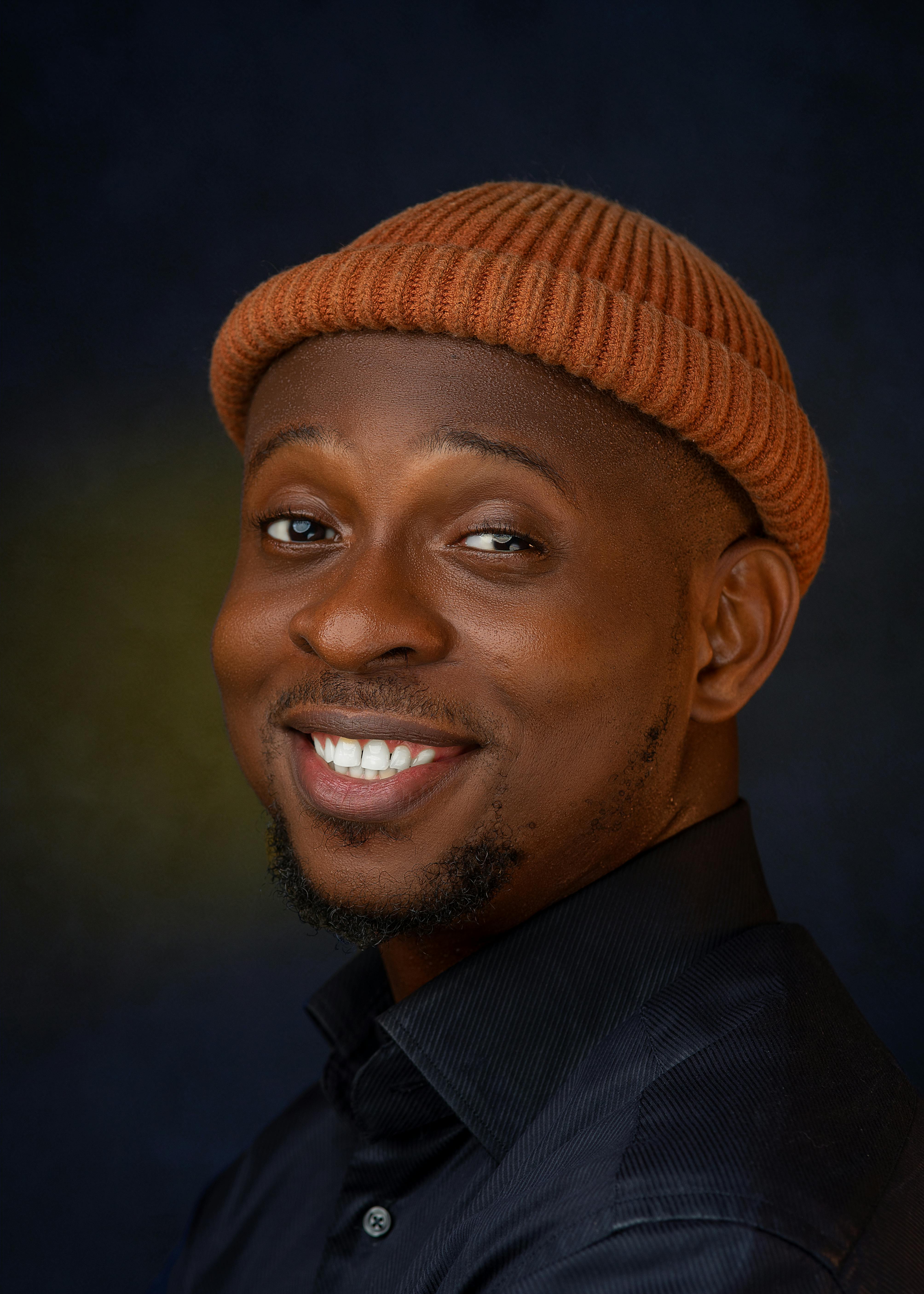 Close-up headshot of a smiling man wearing a brown beanie and black shirt against dark background.