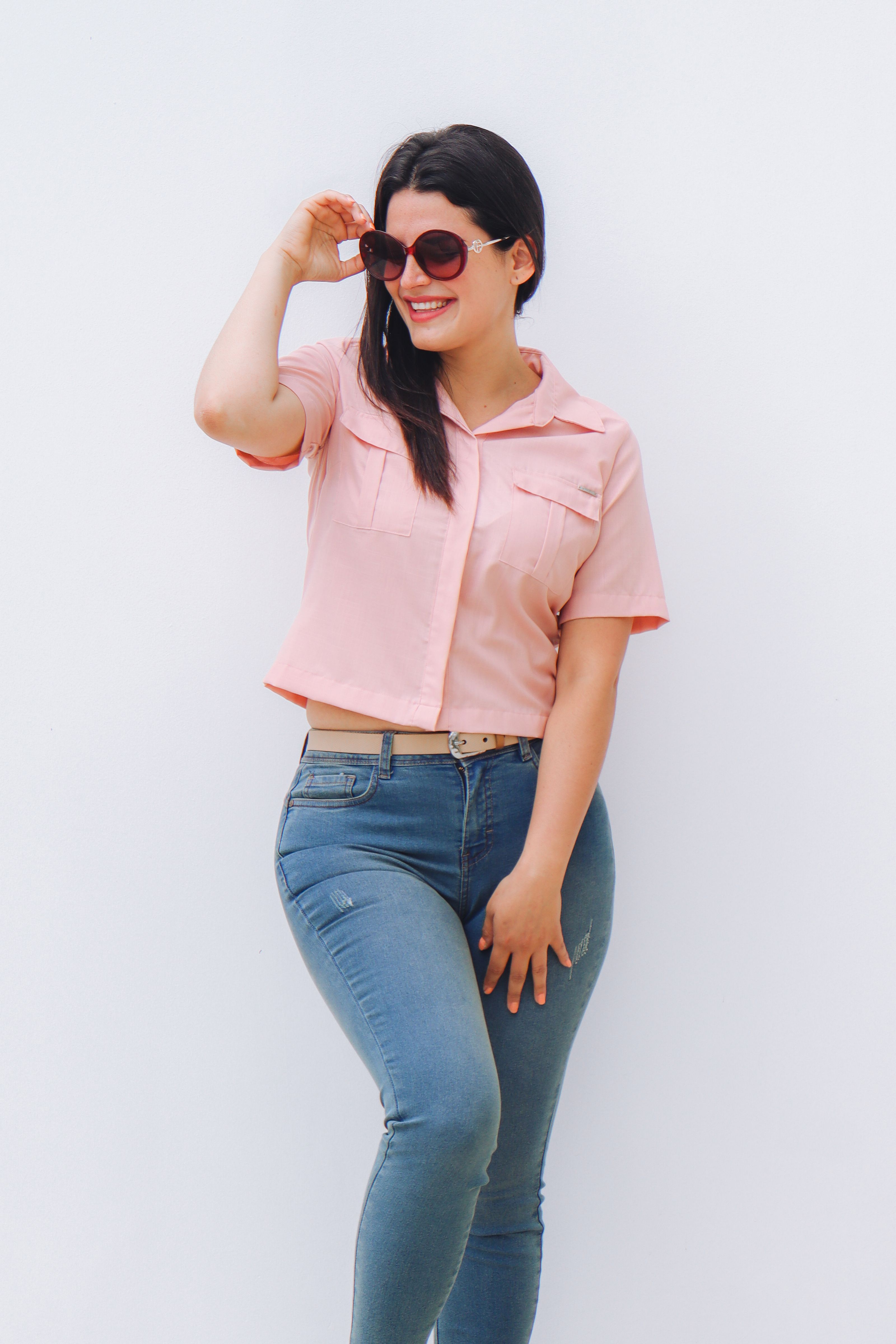 Mulher estilosa em jeans casual e blusa rosa posando com óculos de sol e um sorriso.