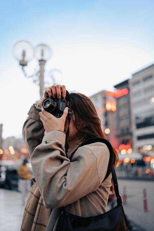 Free A woman in a beige coat captures a city scene with her camera during twilight, showcasing urban life. Stock Photo