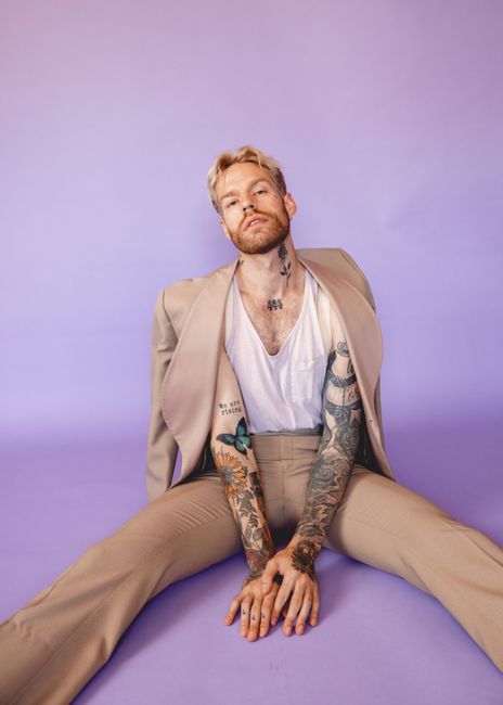 Fashion portrait of a tattooed model in a suit sitting against a purple backdrop.