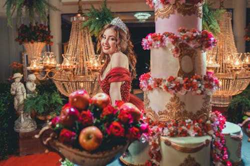 Young Woman in a Dress and a Tiara Standing next to a Table with a Large Cake and Fruit 