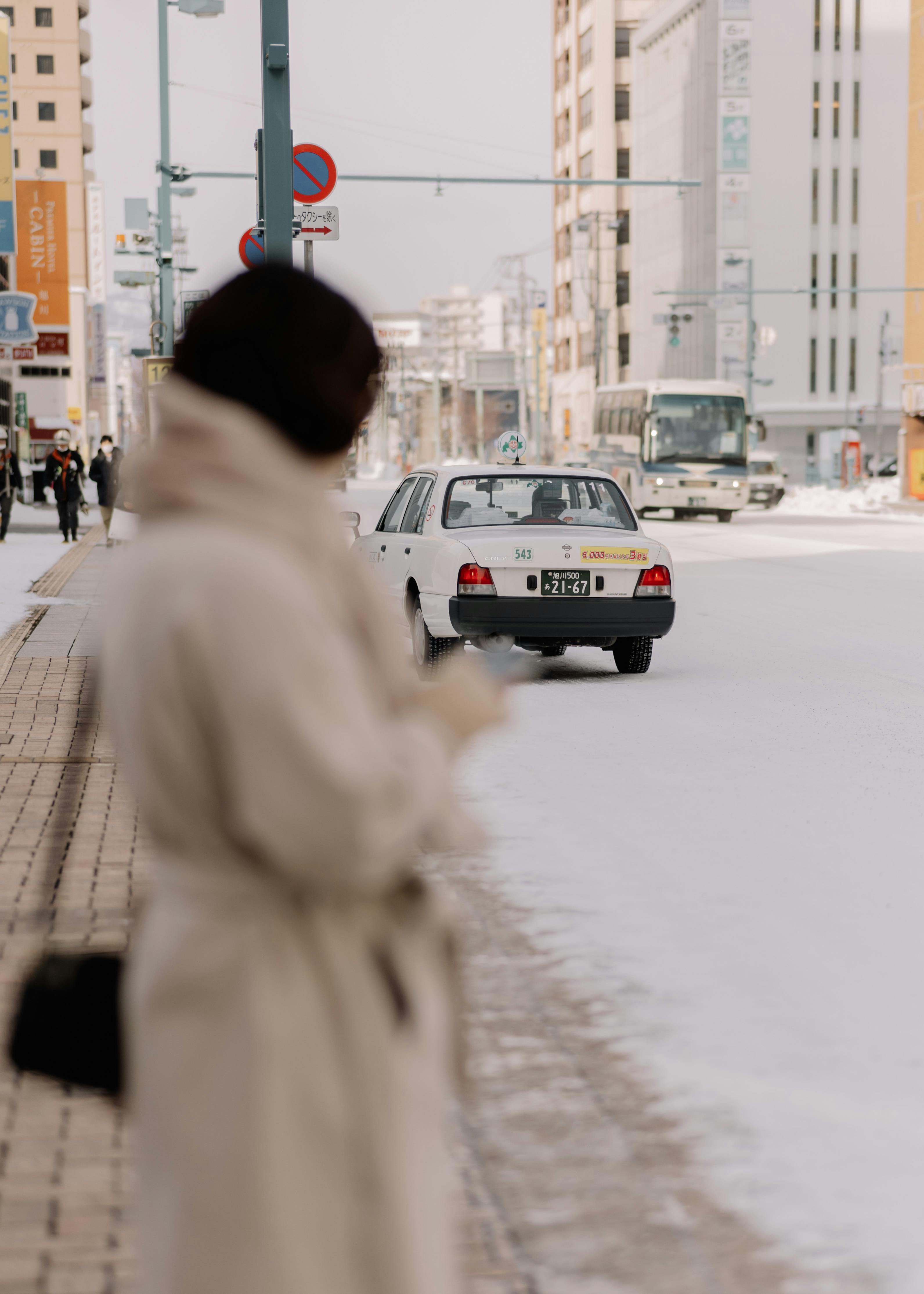 A blurred woman in winter attire stands on a snowy street in Sapporo as traffic passes by.