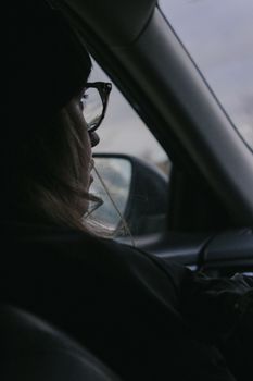 A woman in glasses driving a car, captured from the passenger seat in a close-up view.