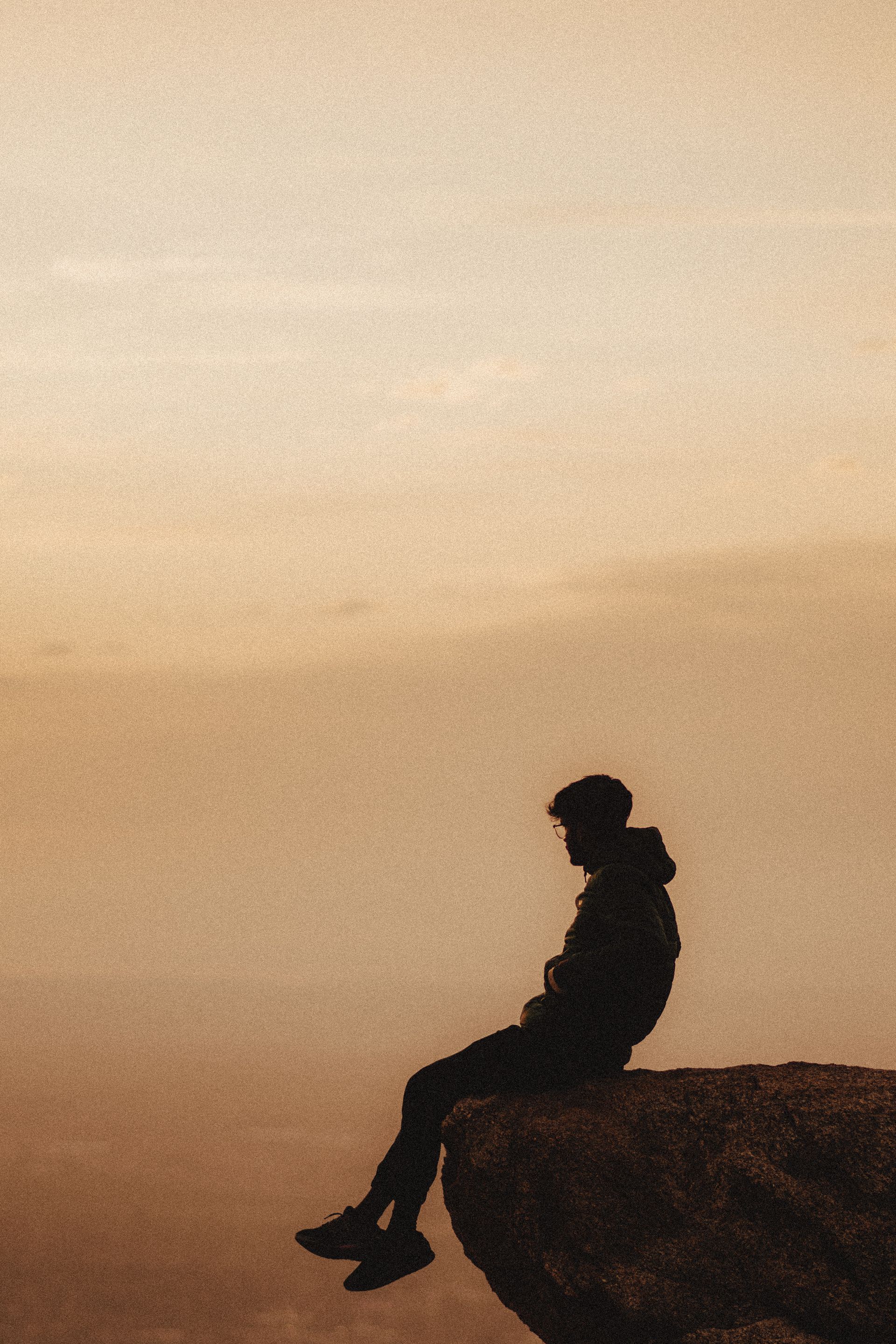 A lone figure seated in stillness at the edge of a cliff as evening falls