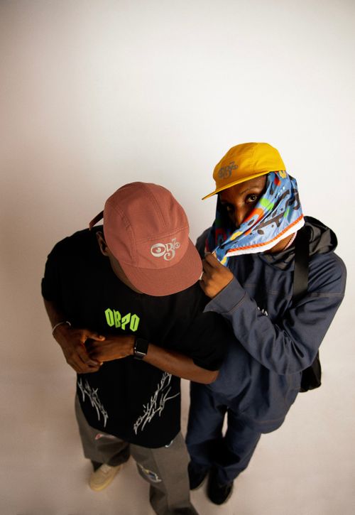 Free Two men in stylish streetwear pose confidently with caps and a colorful shawl against a white background. Stock Photo