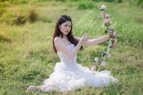 Photo of a Cute Young Woman in a White Dress Sitting on the Grass with a Bow Decorated with Flowers