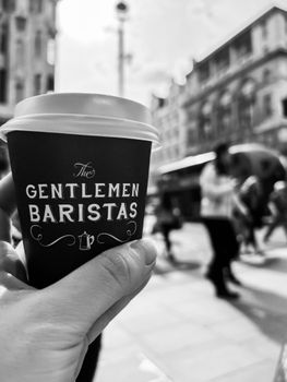 A hand holds a Gentlemen Baristas coffee cup on a bustling city street.