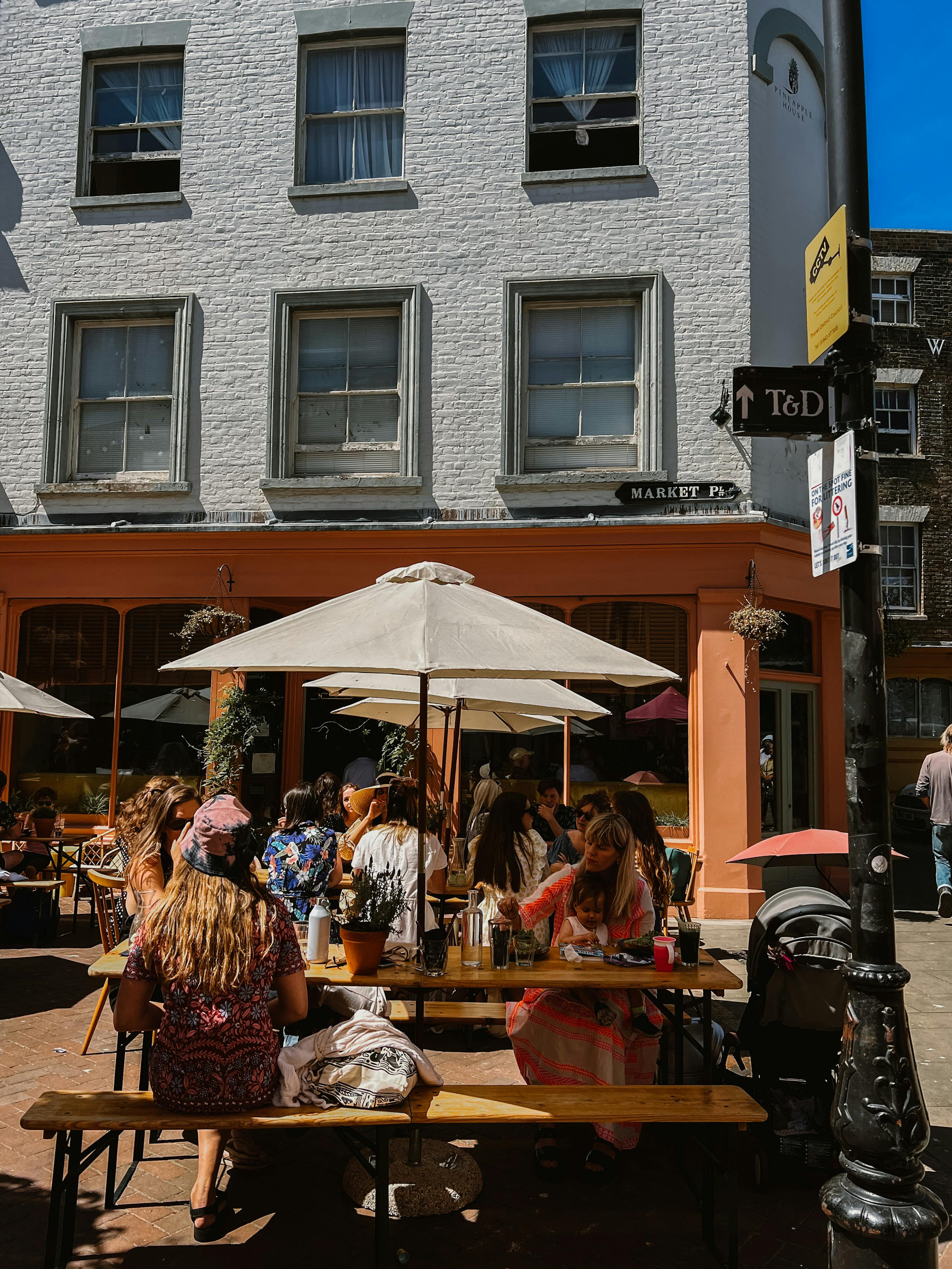 A lively outdoor café with people enjoying a sunny day under umbrellas on a bustling sidewalk.
