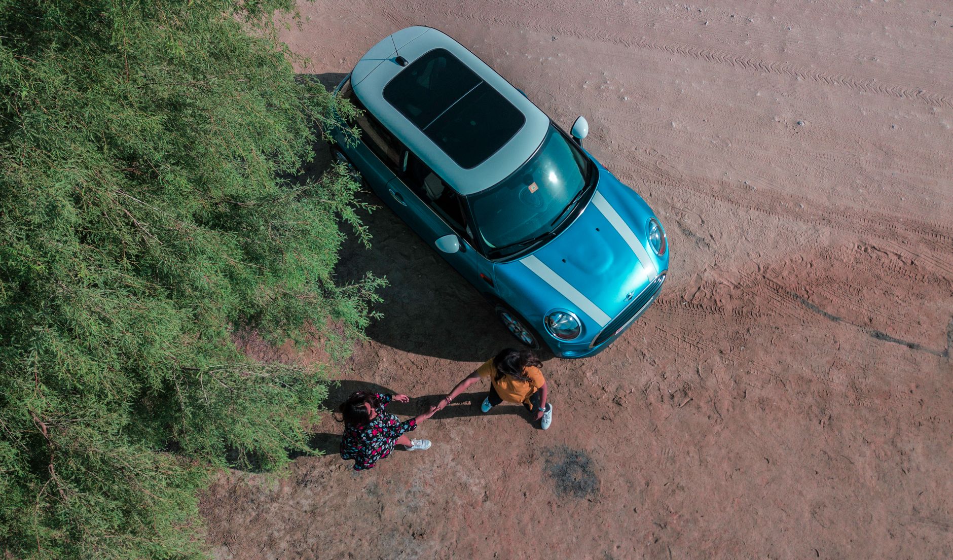 Aerial shot of a blue Mini Cooper on a dirt road with two people standing nearby, surrounded by greenery.