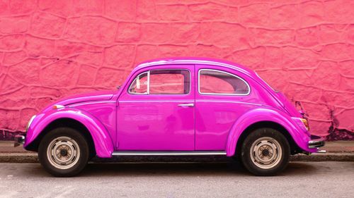 Free Side view of a vibrant pink classic Volkswagen Beetle parked in front of a pink wall in an urban setting. Stock Photo