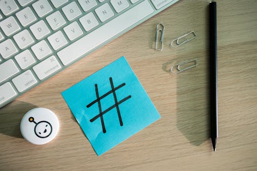 Top View of a Keyboard, Paper Clips, Pencil and a Sticky Note with a Hashtag on It Lying on a Desk 