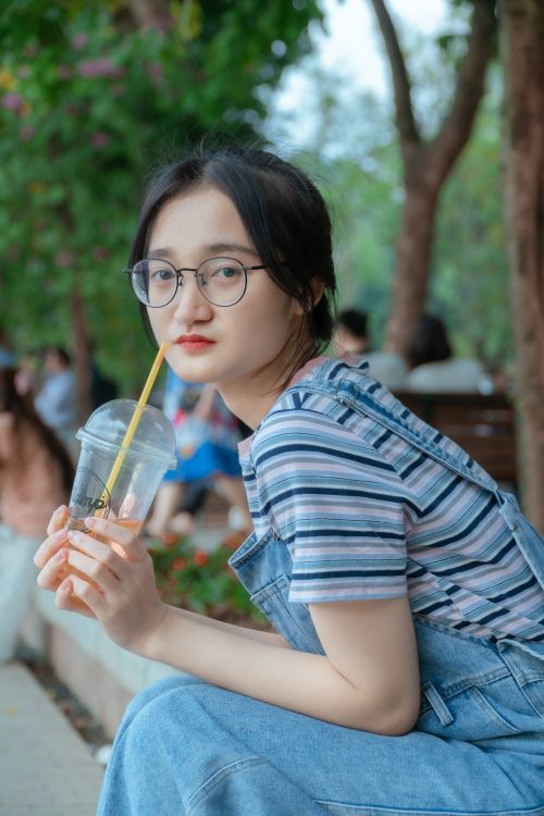 Free A young woman with eyeglasses sits outdoors holding a plastic cup with a straw, surrounded by trees. Stock Photo