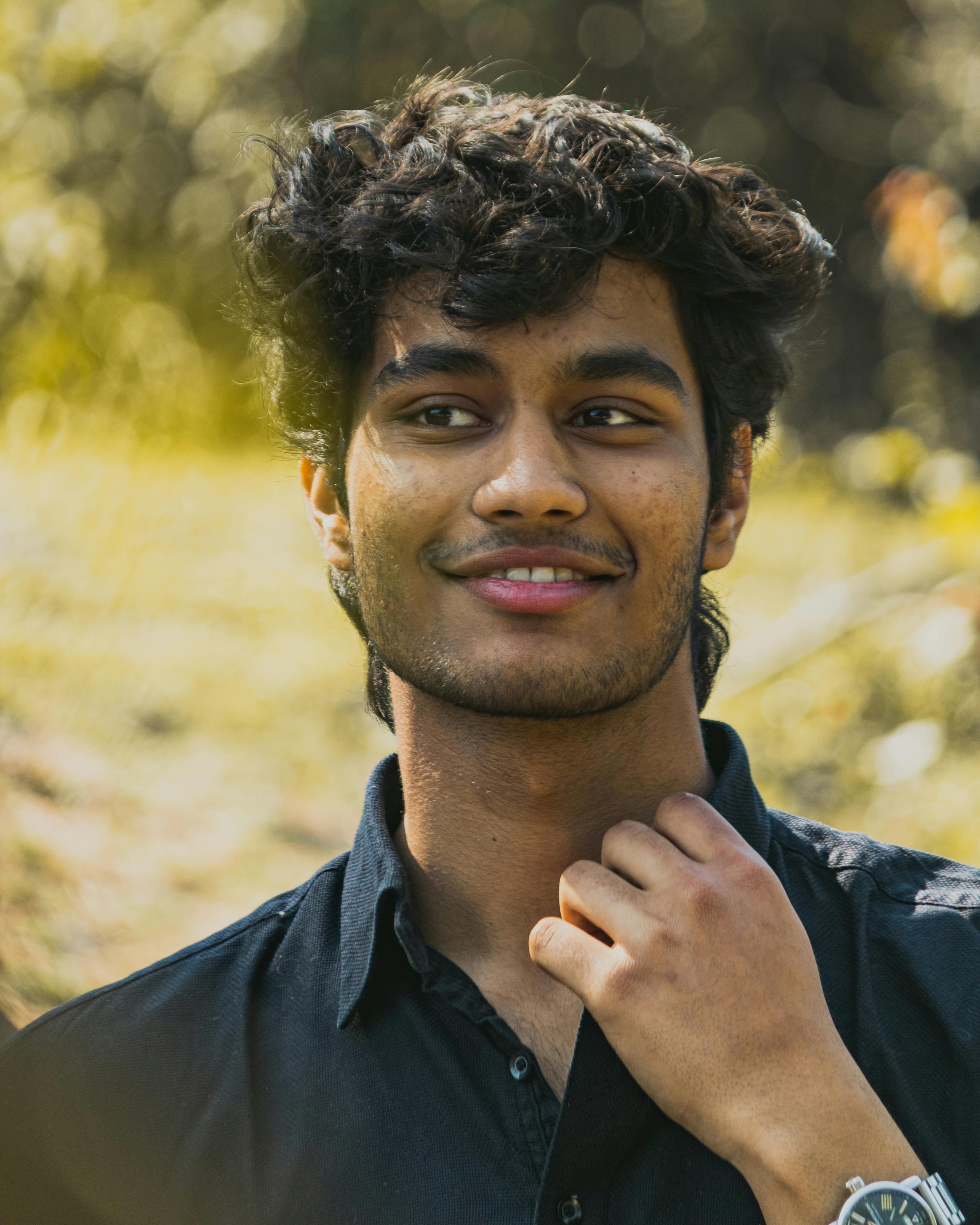 A relaxed young man with a mustache smiles casually outdoors, dressed in a black shirt.