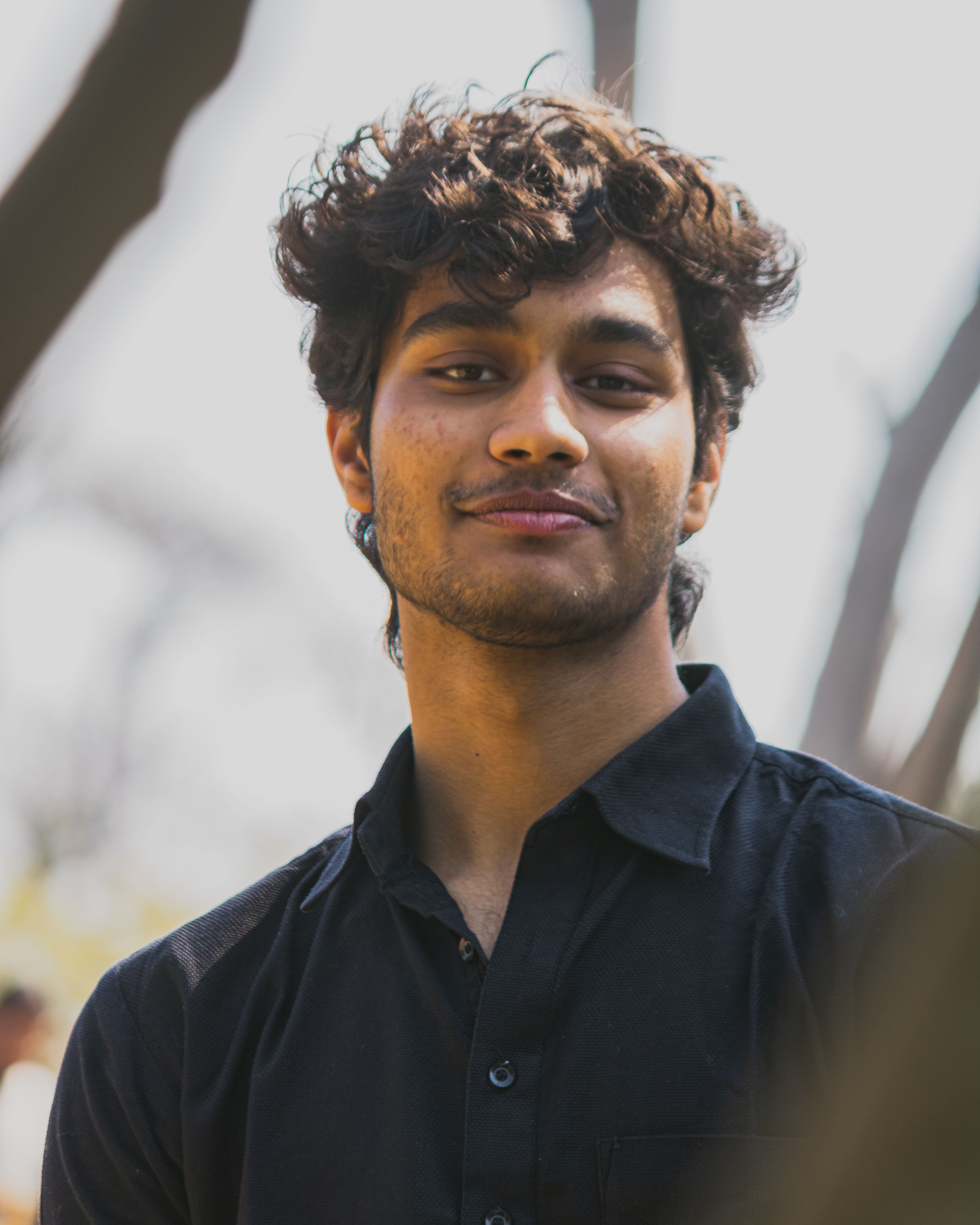 Close-up portrait of a young man with curly hair and a mustache in a natural outdoor setting.