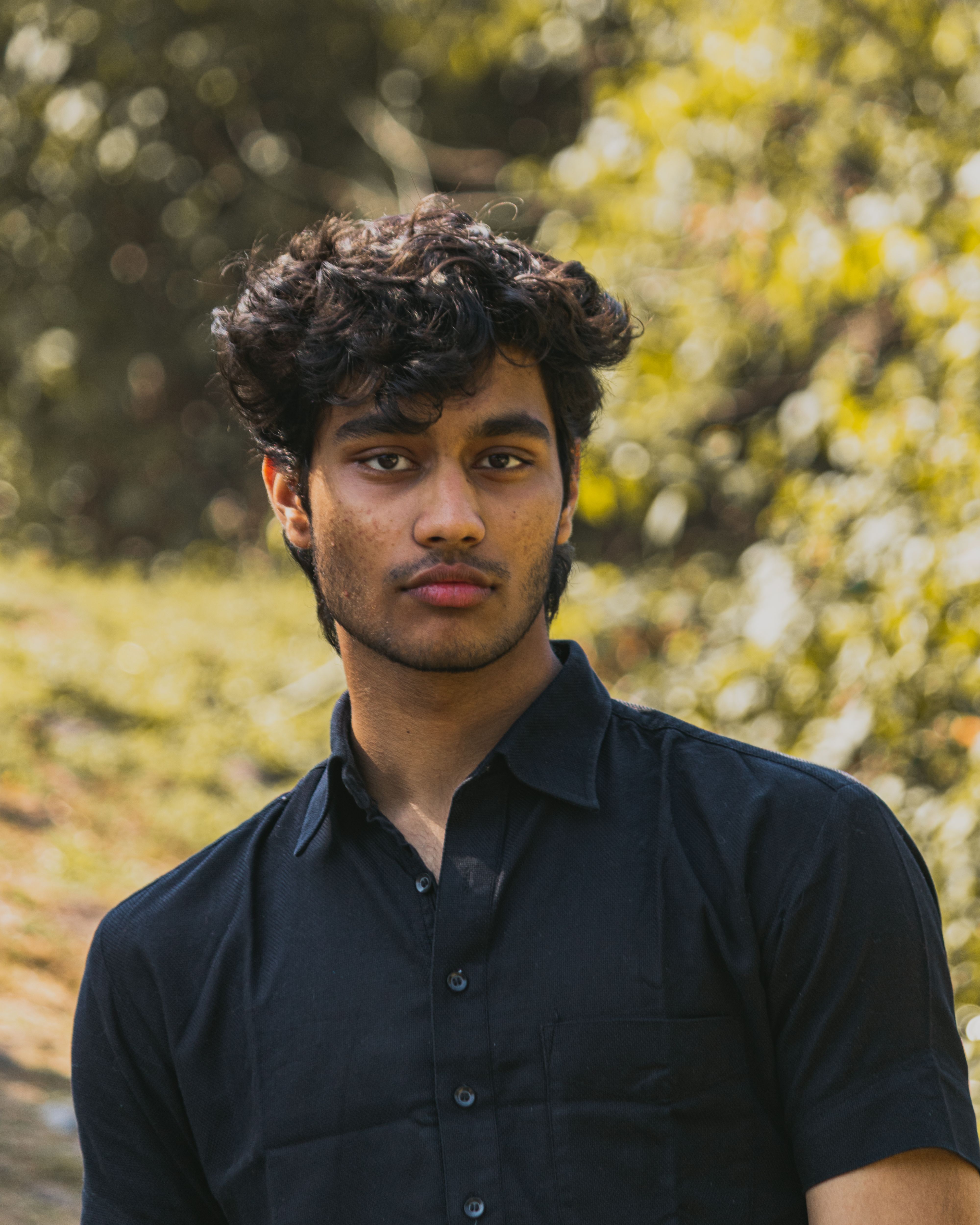 A young man with curly hair wearing a black shirt in a natural outdoor setting.