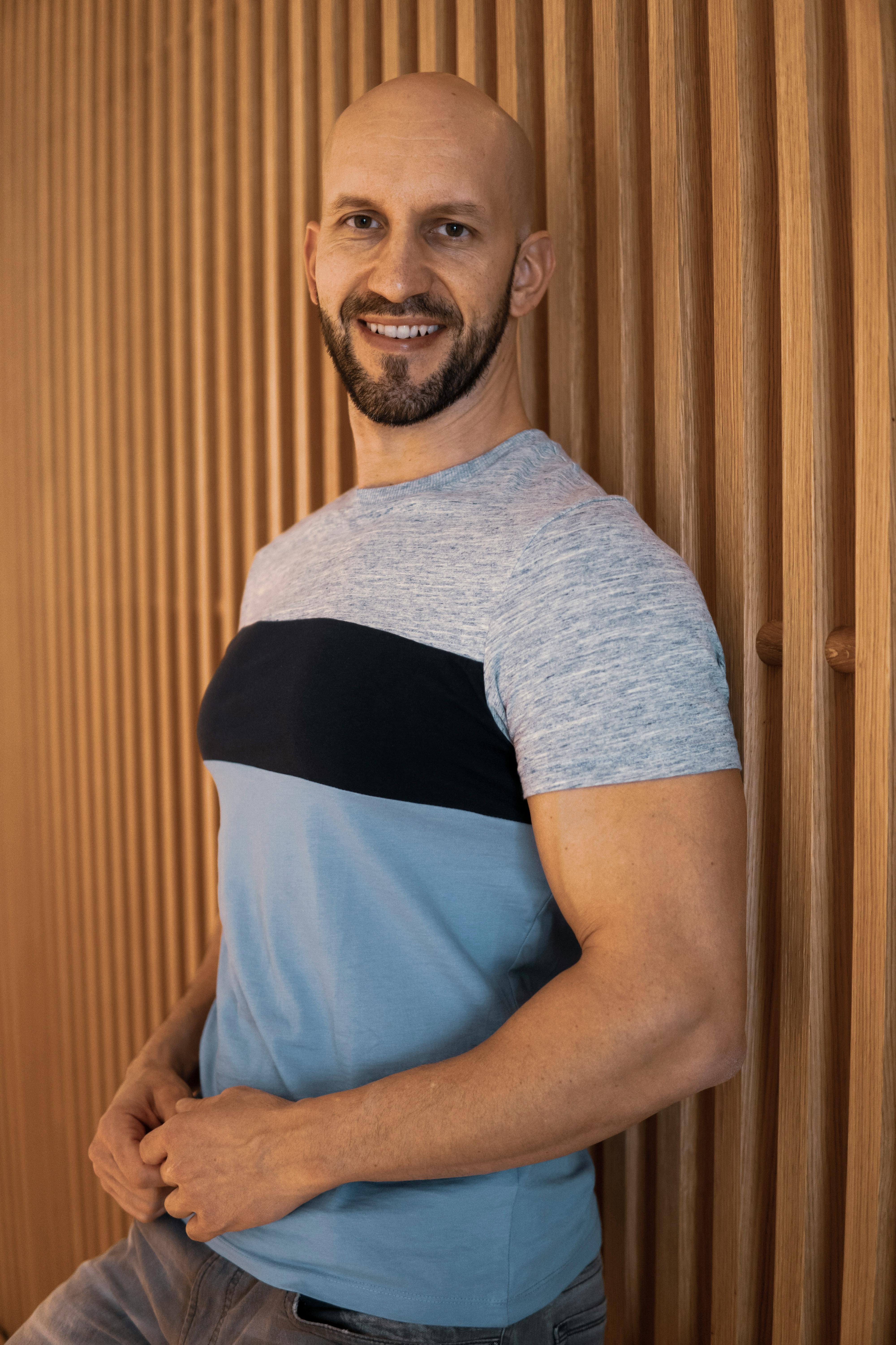 Portrait of a muscular bald man smiling indoors against a wooden background.