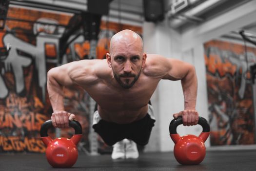 Muscular bald man doing push ups with kettlebells in gym.