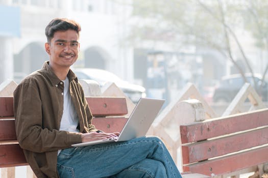 Young man sitting on a bench outdoors using a laptop, smiling at the camera.