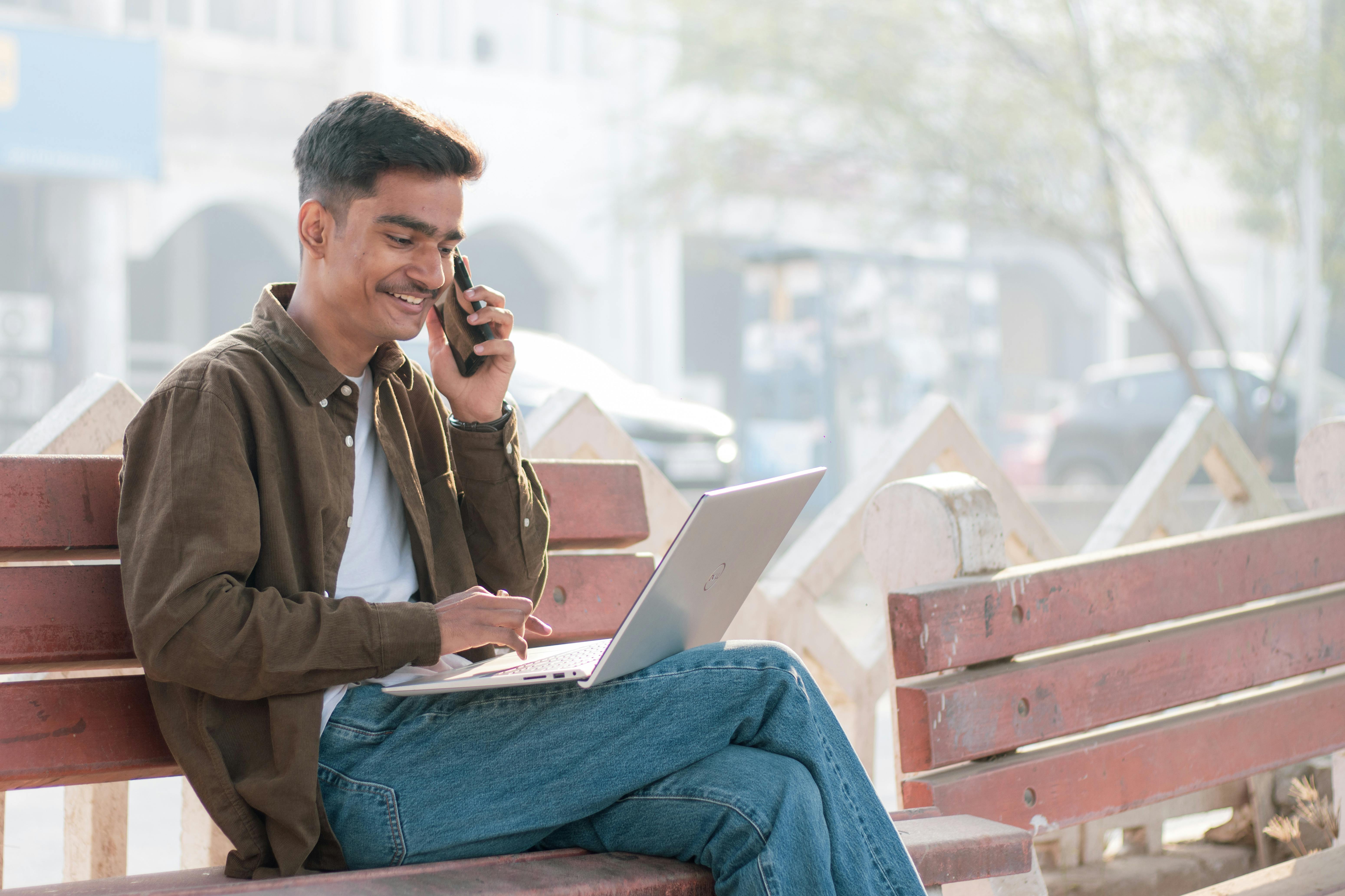 Young man working remotely outdoors with laptop and phone, enjoying productivity on a sunny day.