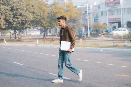 Free Casual young man crosses a city street holding a laptop, showcasing urban lifestyle. Stock Photo