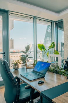 A bright modern home office with large windows, plants, and a laptop on a desk.
