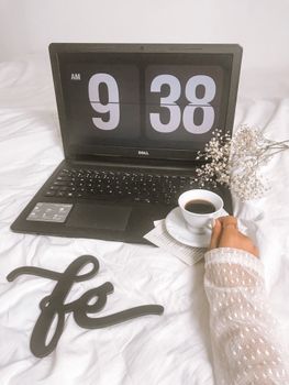 Comfortable morning setup showing a coffee cup and laptop displaying 9:38 AM on white bedding.