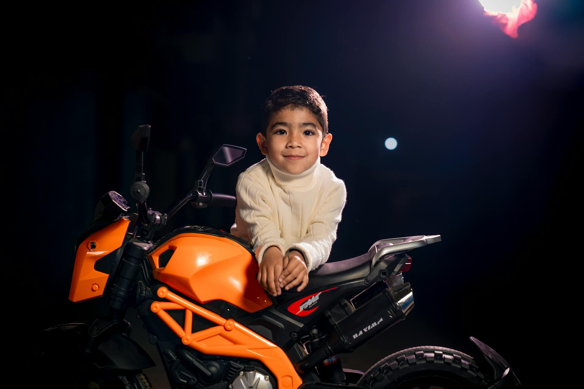 Smiling boy in a sweater sitting on a vibrant mini motorcycle under night lighting.