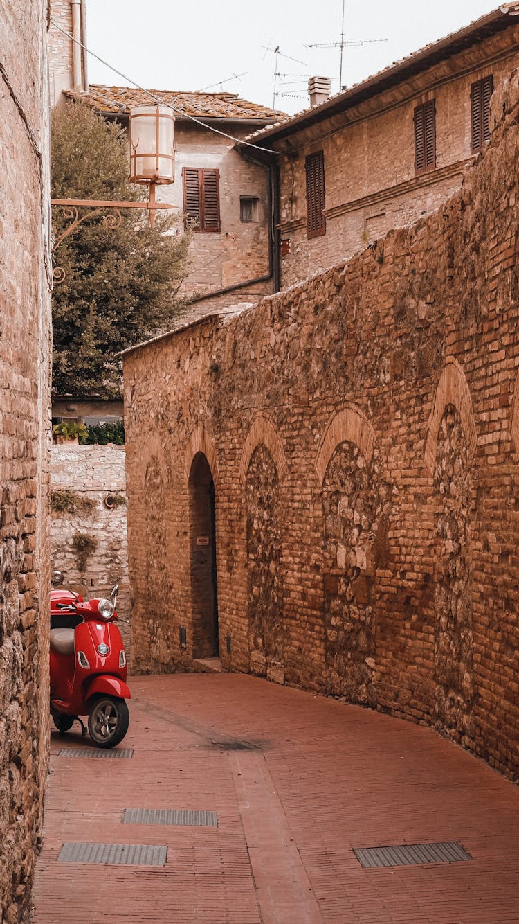 A Red Motor Scooter Parked In A Narrow Alley In The Town Of San Gimignano, Tuscany, Italy 