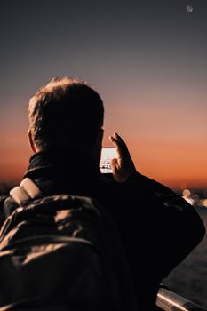 A man with a backpack captures a stunning sunset view using his smartphone as the sky transitions to twilight.