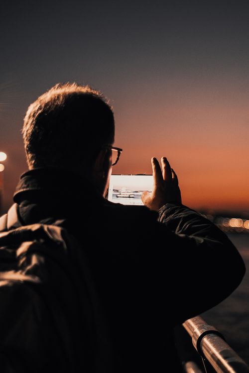 Free A man with a backpack takes a photo of the sunset over water with a smartphone from a pier. Stock Photo