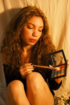 A young woman applies eye shadow from a colorful palette with a soft makeup brush.
