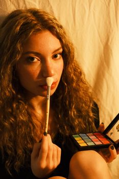 Caucasian woman with curly hair applying makeup with a brush.