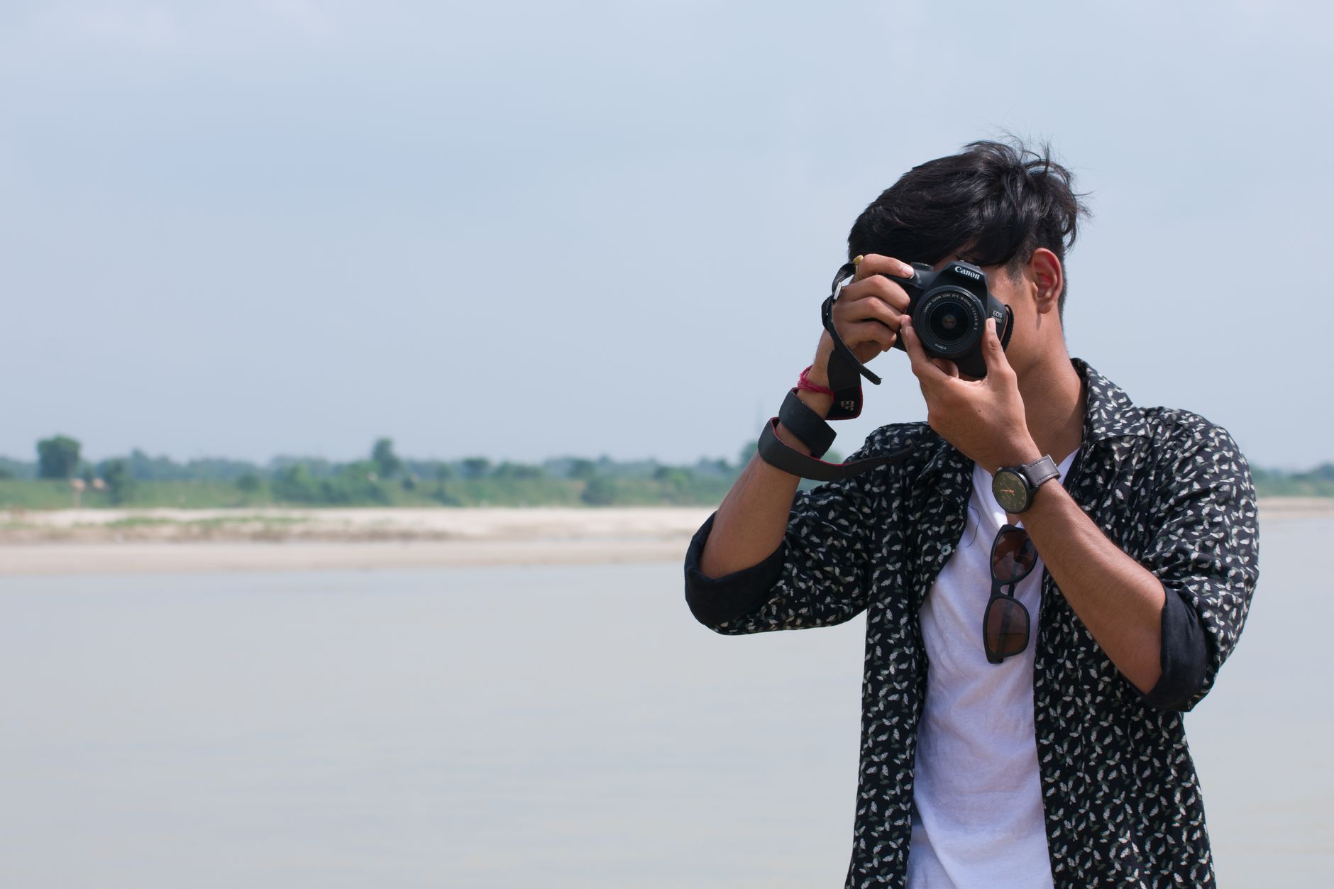 A young man with a DSLR camera photographing near a lakeside on a sunny day.