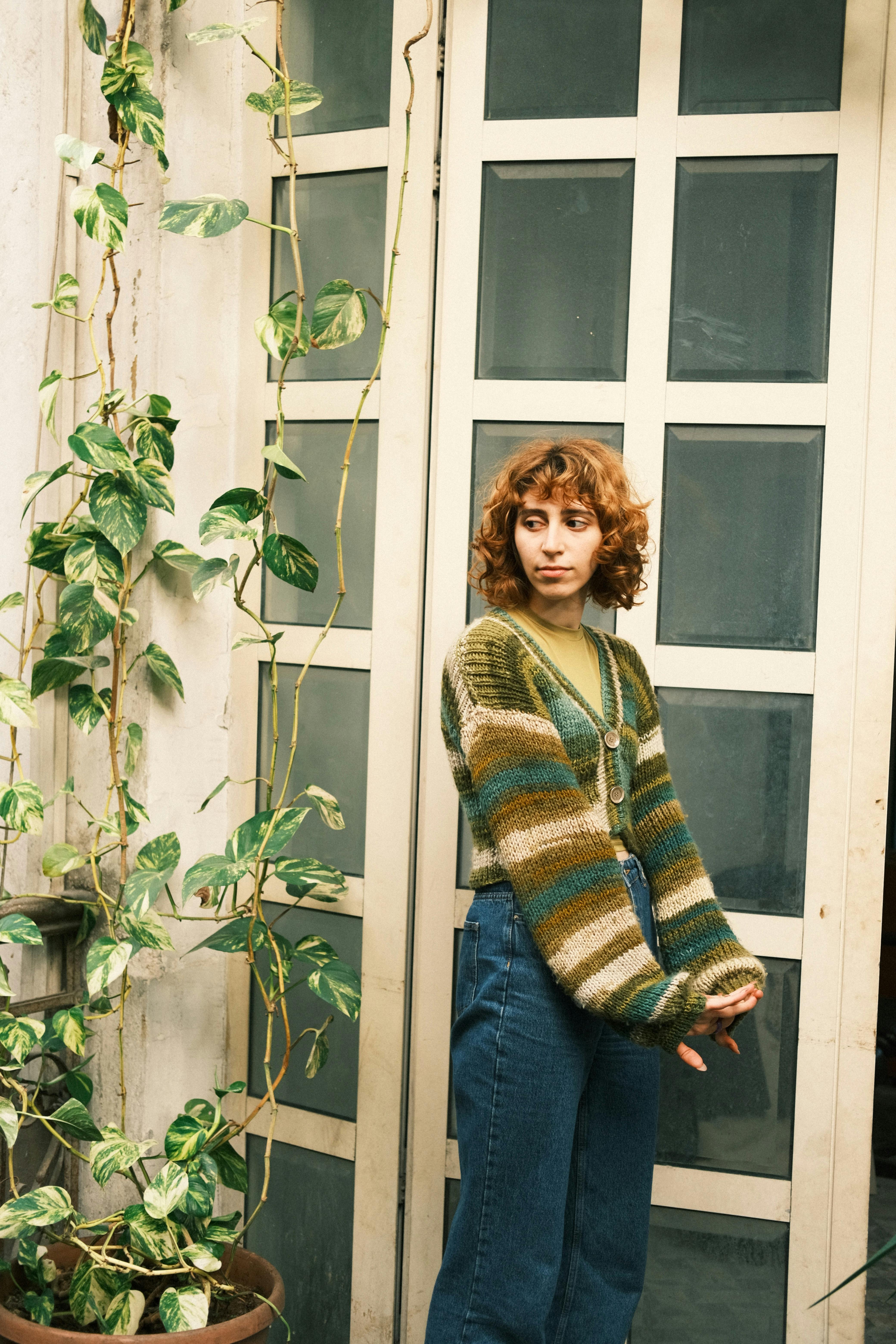 Young woman in a trendy sweater stands by a doorway with greenery.