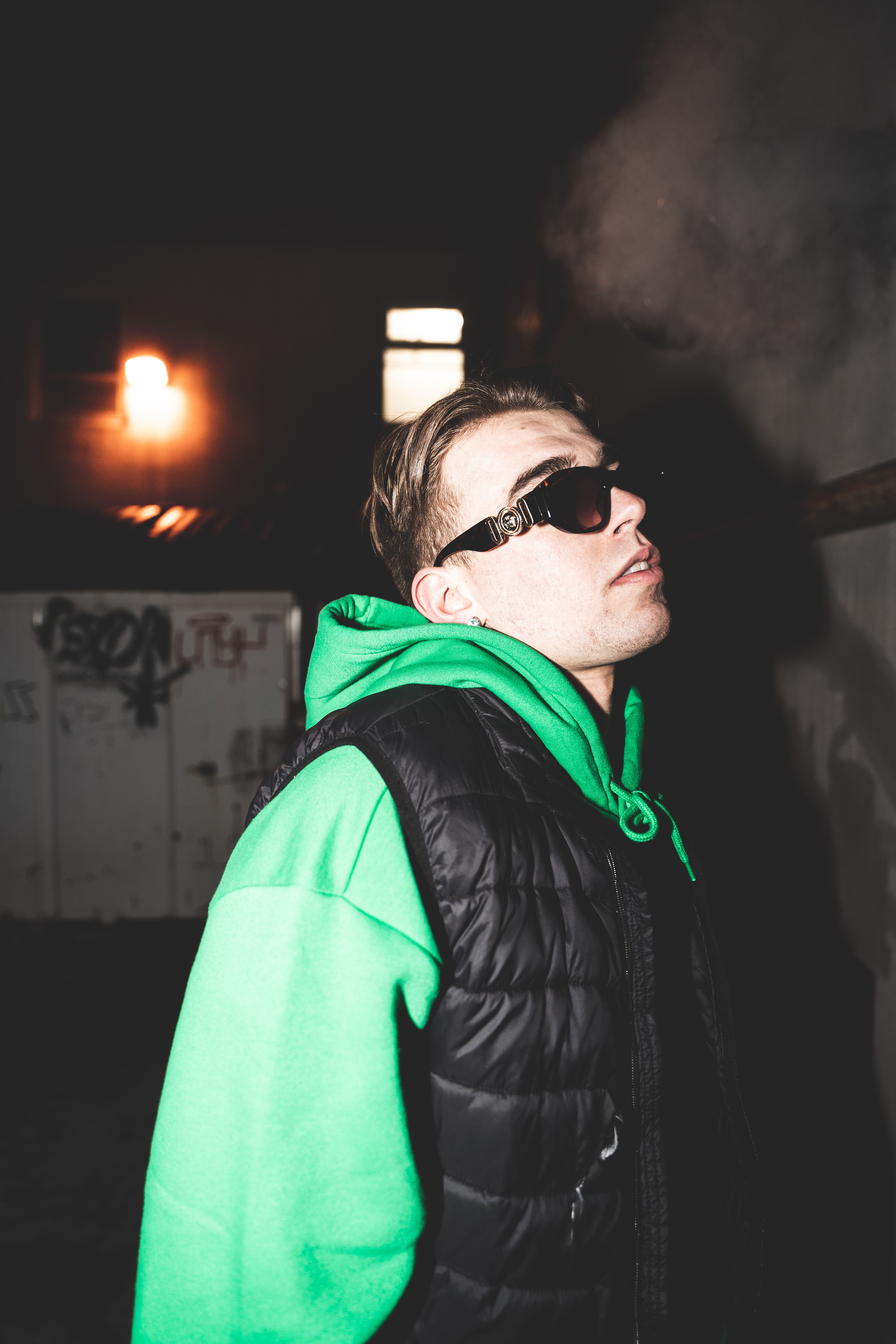 Portrait of a man wearing a green hoodie and sunglasses at night with smoke in the background.