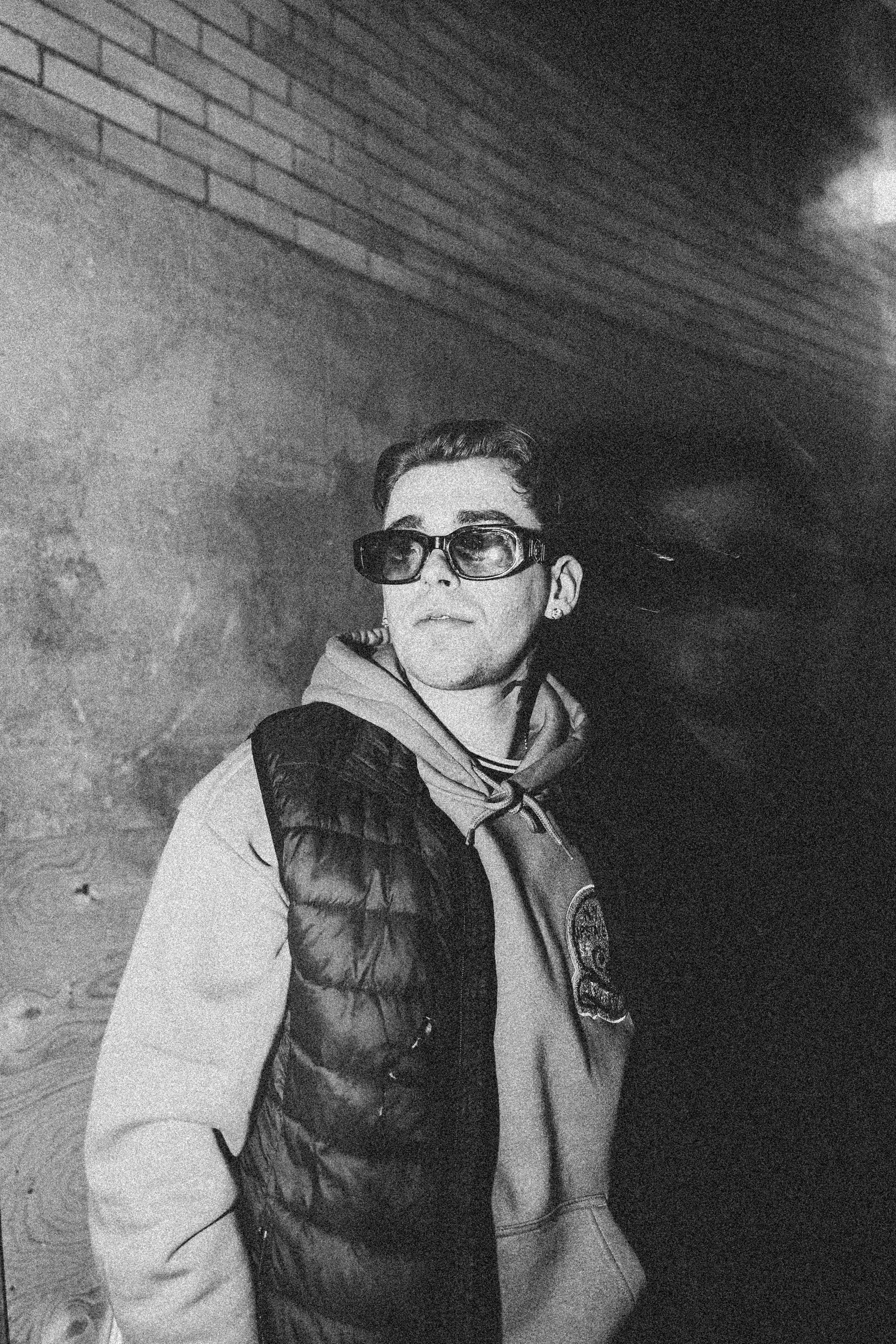 Black and white portrait of a man in sunglasses, jacket, and hoodie against an urban backdrop.