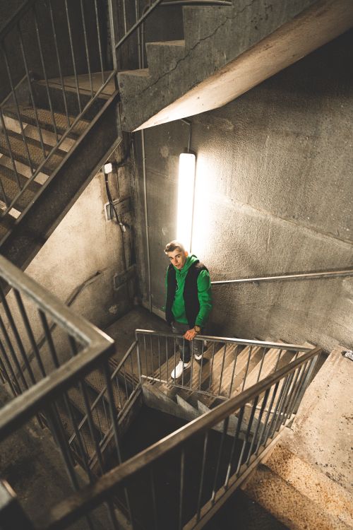 Free A man in a green hoodie stands on a concrete staircase, lit by modern lighting. Stock Photo