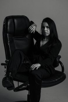 A confident woman in a dark outfit seated on an office chair, showcasing modern fashion in a studio setting.