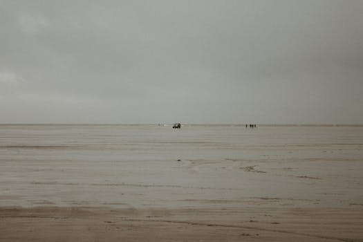 A tranquil scene of a car and people on an expansive, empty beach under a cloudy sky.